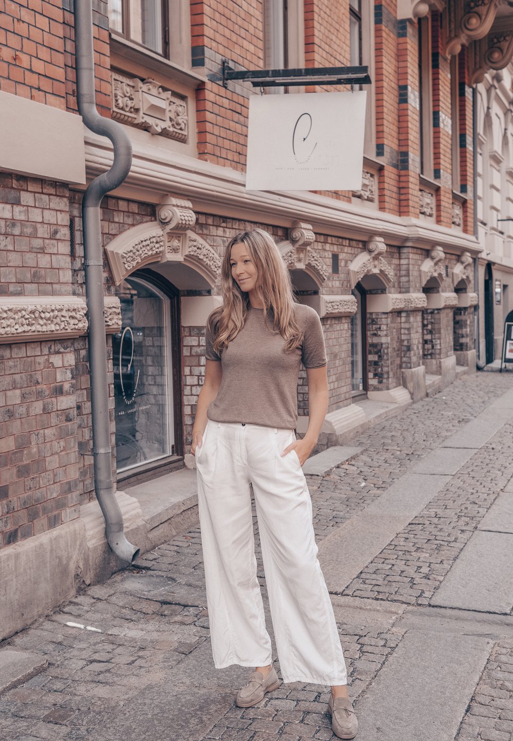 Beige kortärmad topp i kombination med vita vida byxor med sidofickor, kompletterat med ljusgrå skor, mot en tegelvägg.