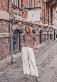 Beige kortärmad topp i kombination med vita vida byxor med sidofickor, kompletterat med ljusgrå skor, mot en tegelvägg.