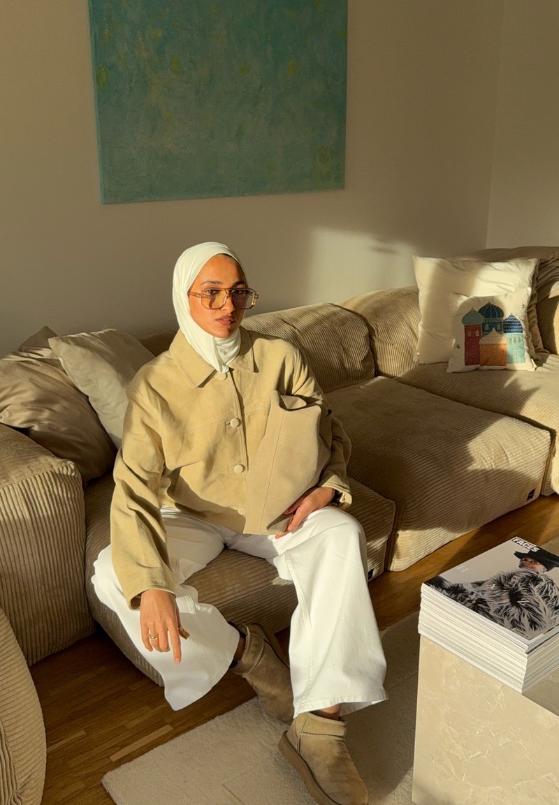 Veste beige surdimensionnée à boutons, pantalon blanc et bottines marron. Assis sur un canapé rayé avec un coussin décoratif et des magazines.