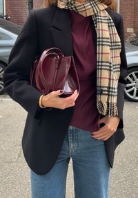 Blazer nero, top bordeaux e jeans a vita alta blu. Tiene una piccola borsa bordeaux con finitura lucida e una sciarpa beige a quadri.