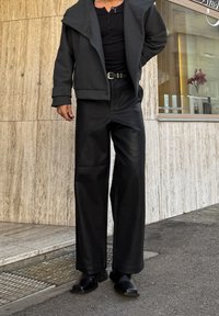 Personne portant un sweat à capuche sombre, une chemise noire à boutons, un pantalon large en cuir noir, une ceinture et des bottes noires, debout sur un trottoir en ville près d'un mur en pierre.