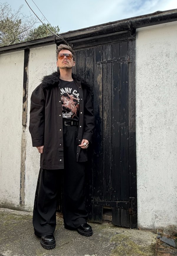 Bărbat purtând ochelari de soare maro, palton negru cu guler de blană, tricou grafic, pantaloni negri largi și pantofi cu platformă, stând lângă o ușă neagră uzată.