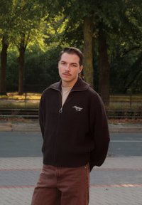Brauner Reißverschluss-Strickpullover mit Stehkragen und Logo, kombiniert mit hellbraunen Hosen. Der Stoff wirkt strukturiert, vor einem natürlichen Hintergrund.