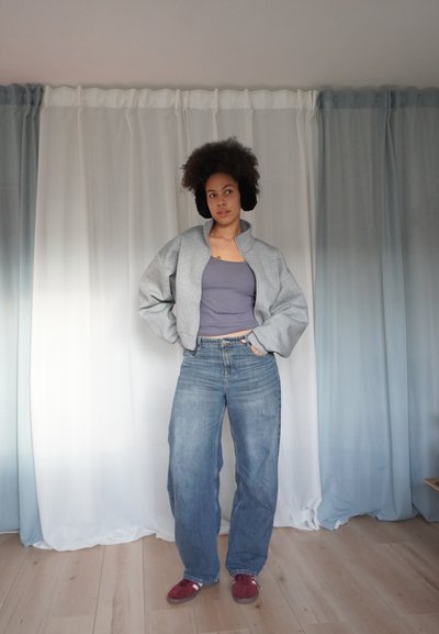 Cardigan gris corto sobre una camiseta sin mangas púrpura, combinado con vaqueros de pierna ancha en azul claro y zapatillas rojas. Lleva orejeras negras y esponjosas.