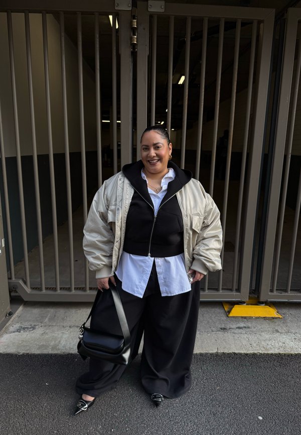 Kobieta stojąca przed dużą metalową bramą, ubrana w warstwową czarno-białą stylizację z beżową kurtką puchową, trzymająca czarną torebkę, uśmiechająca się.