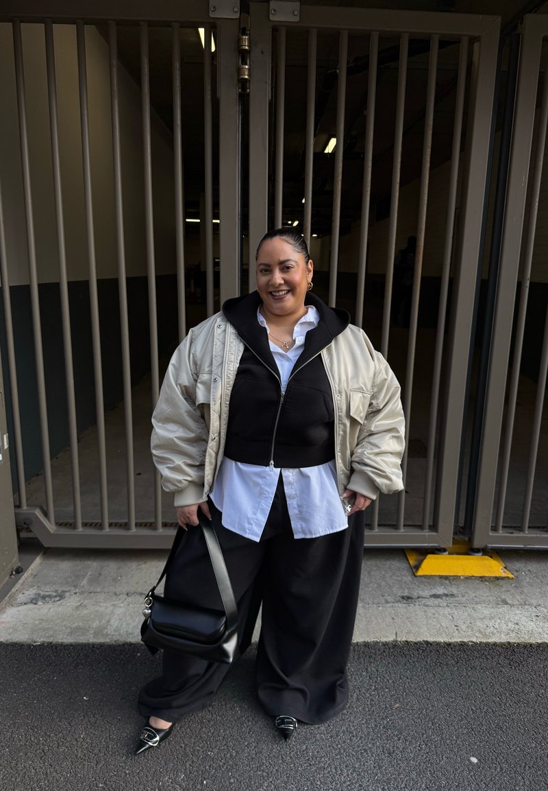 Femme debout devant une grande porte métallique, vêtue d'une tenue noire et blanche superposée avec une doudoune beige, tenant un sac à main noir, souriante.