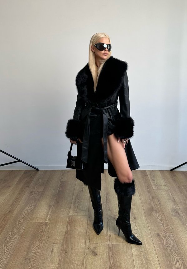 Woman with long blonde hair wearing oversized black sunglasses, a black coat trimmed with fur, high-heeled boots, and holding a small black handbag.