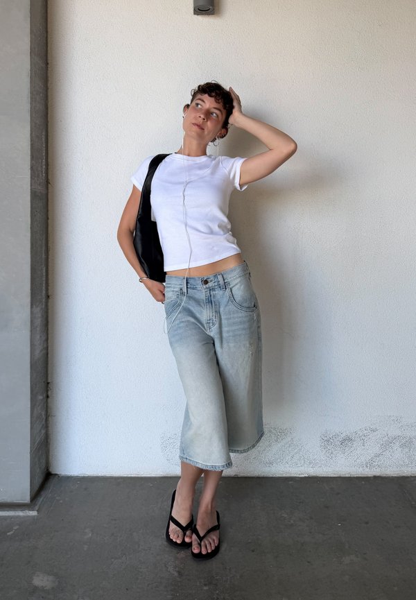 Žena s kratkom kovrčavom kosom koja nosi bijelu majicu, traper culottes hlače, crne sandale, slušalice i crnu torbu na rame, naslonjena na bijeli zid.
