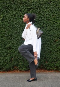 Czarna torebka na złotym łańcuszku, biała koszula na guziki, czarne dżinsy i czarne baleriny. Stoi na tle zielonego krzewu.