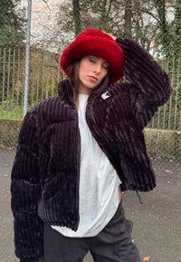 Personne portant un chapeau rouge en peluche, une veste noire côtelée, une chemise blanche et un pantalon noir, debout à l'extérieur près d'une clôture en grillage.