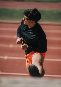 Athlet in schwarzem Langarmshirt und orangefarbene Shorts, der draußen sein Bein auf einer roten Laufbahn dehnt.