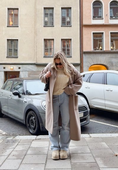 Abrigo largo beige sobre un suéter de punto trenzado color crema, jeans azul claro y zapatos beige de tipo slip-on; bolso negro; de pie en un entorno urbano.