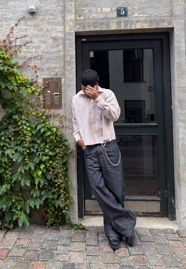 Bežný outfit pozostávajúci zo svetloružovej kockovanej košele na zapínanie a tmavomodrých džínsov s širokými nohavicami a strieborným reťazovým detailom. Čierna čiapka a topánky.