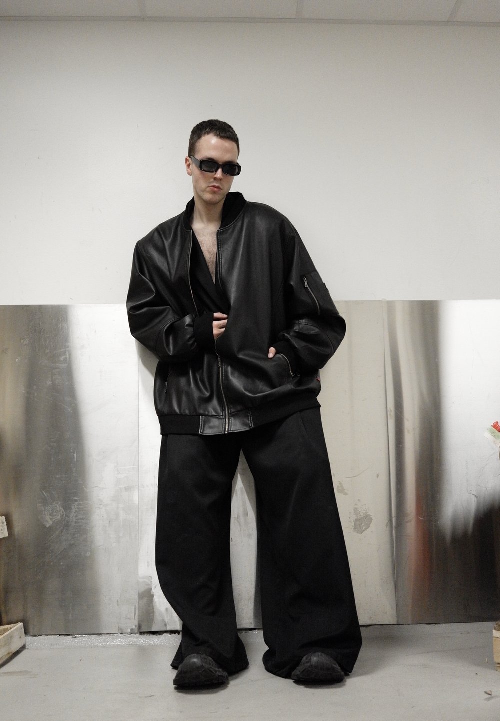 Veste bombardier en cuir noir à coupe oversize, manches côtelées et fermeture éclair, associée à un pantalon large noir et des chaussures noires. Lunettes de soleil foncées portées.