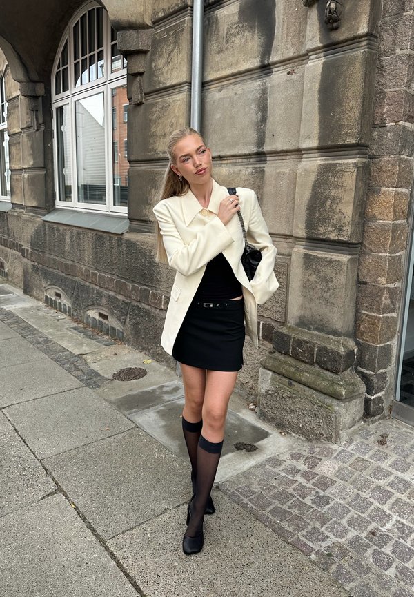 Una mujer con una americana blanca, una falda negra y calcetines transparentes hasta la rodilla está de pie junto a un edificio de piedra, sujetando la correa de su bolso de hombro.