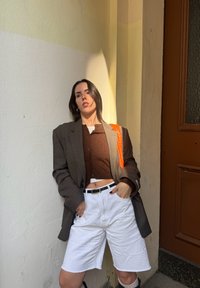 Brun struktureret blazer over en brun cropped sweater, hvide shorts, sort bælte og taske med orange accenter. Står mod en lys væg.