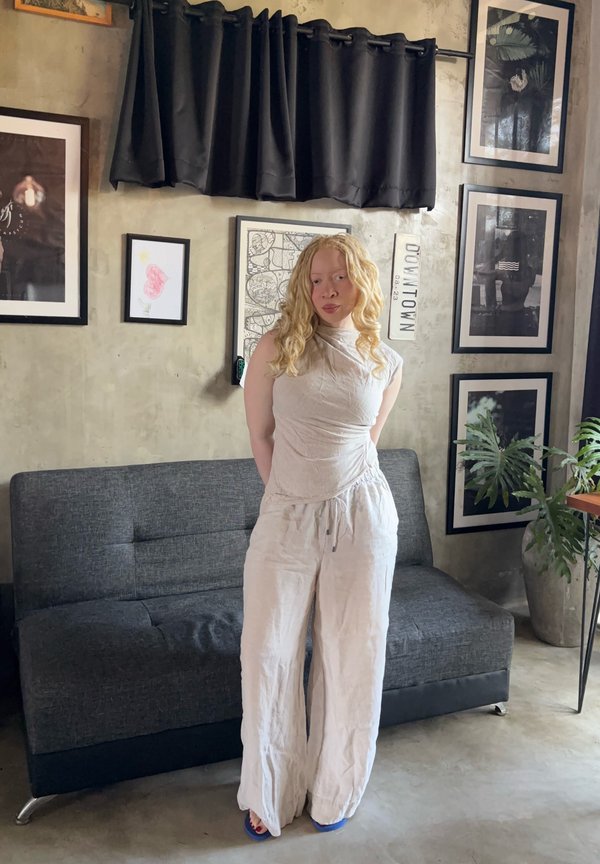 Femme aux longs cheveux blonds bouclés portant une tenue blanche, debout devant un canapé gris dans une pièce avec des œuvres encadrées au mur et des plantes d’intérieur.