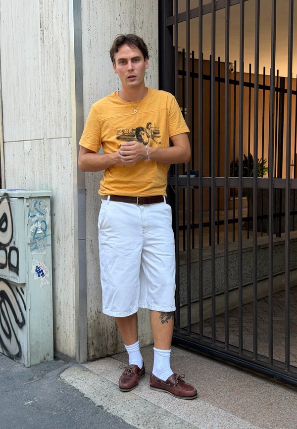 Jonge man staat met gevouwen handen, draagt een geel grafisch t-shirt, witte short, witte sokken en bruine schoenen bij een metalen poort.