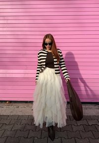 Cardigan rayé noir et blanc porté sur un haut marron, jupe en tulle blanche superposée, bottines noires pointues, et sac marron devant un mur rose.