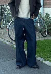 Traperice širokih nogavica tamne boje s visokim strukom i opuštenim krojem. Uparene s laganim pamučnim džemperom i crnim cipelama, u pozadini se vide bicikli.