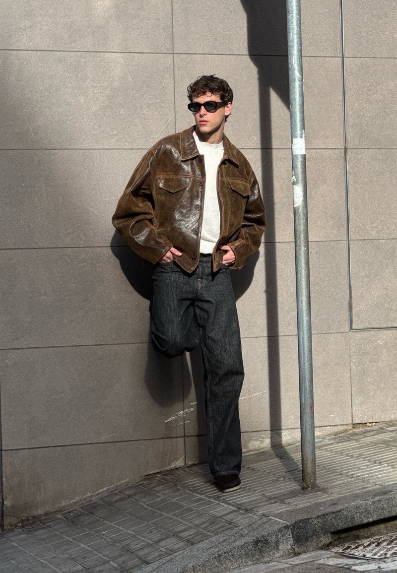 Brown leather jacket with two chest pockets, cream jumper, and dark denim trousers, set against a grey wall, with a lamppost nearby.
