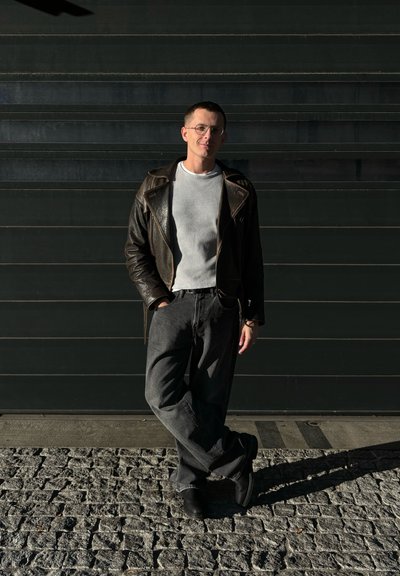 Chaqueta de cuero negra tipo biker sobre una camiseta de canalé gris claro, combinada con unos pantalones de pierna ancha gris oscuro y zapatos negros, sobre una superficie de piedra texturizada.