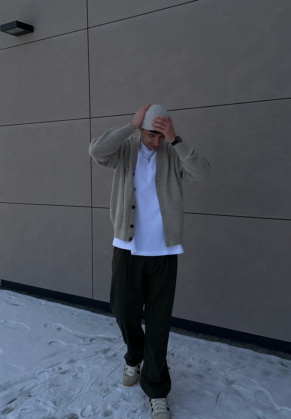 Cardigan en maille gris clair, t-shirt blanc, pantalon vert olive et un bonnet léger. Le cardigan est orné de boutons, la personne est debout sur la neige.