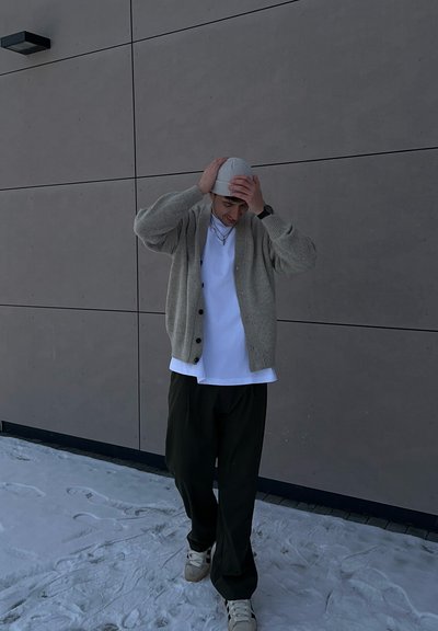 Cárdigan de punto gris claro, camiseta blanca, pantalones verde oliva y un gorro ligero. Presenta botones en el cárdigan, está de pie sobre la nieve.