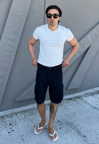 Hombre con gafas de sol negras, camiseta blanca, pantalones cortos cargo negros y chanclas blancas se está ajustando los pantalones contra una pared texturizada gris.