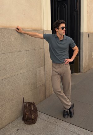 Hombre con gafas de sol, camisa gris y pantalones beige apoyado contra una pared de piedra con una bolsa marrón en el suelo cerca.