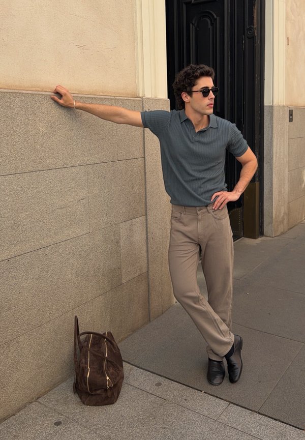 Homme portant des lunettes de soleil, une chemise grise et un pantalon beige appuyé contre un mur de pierre avec un sac marron posé par terre à proximité.