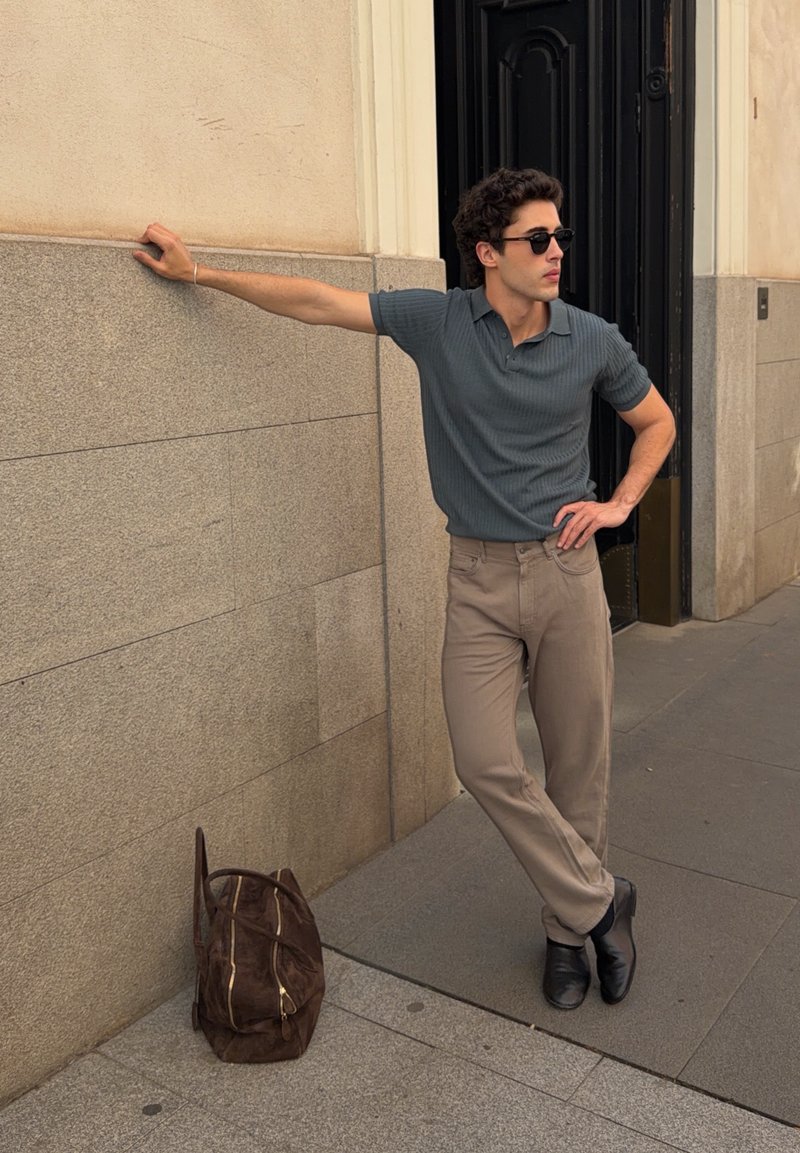 Homme portant des lunettes de soleil, une chemise grise et un pantalon beige appuyé contre un mur en pierre avec un sac marron posé sur le sol à proximité.