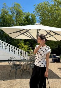 Biała koszula w czarne kropki, ze swobodnym krojem, noszona z czarnymi spodniami o szerokich nogawkach. Tło przedstawia zieleń i meble ogrodowe pod parasolem.