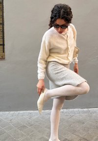 Valge pehme kardigan nuppudega, paaritatud halli põlveni ulatuva seelikuga. Nahatooniga soonikkoes sukkpüksid ja teravate varvastega valkjashallid kontsakingad.