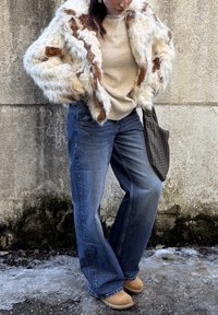 Femme portant une veste en fausse fourrure blanche et marron, un pull beige, un jean bleu ample, des chaussures marron clair, et portant un sac cabas tissé foncé contre un mur en béton.