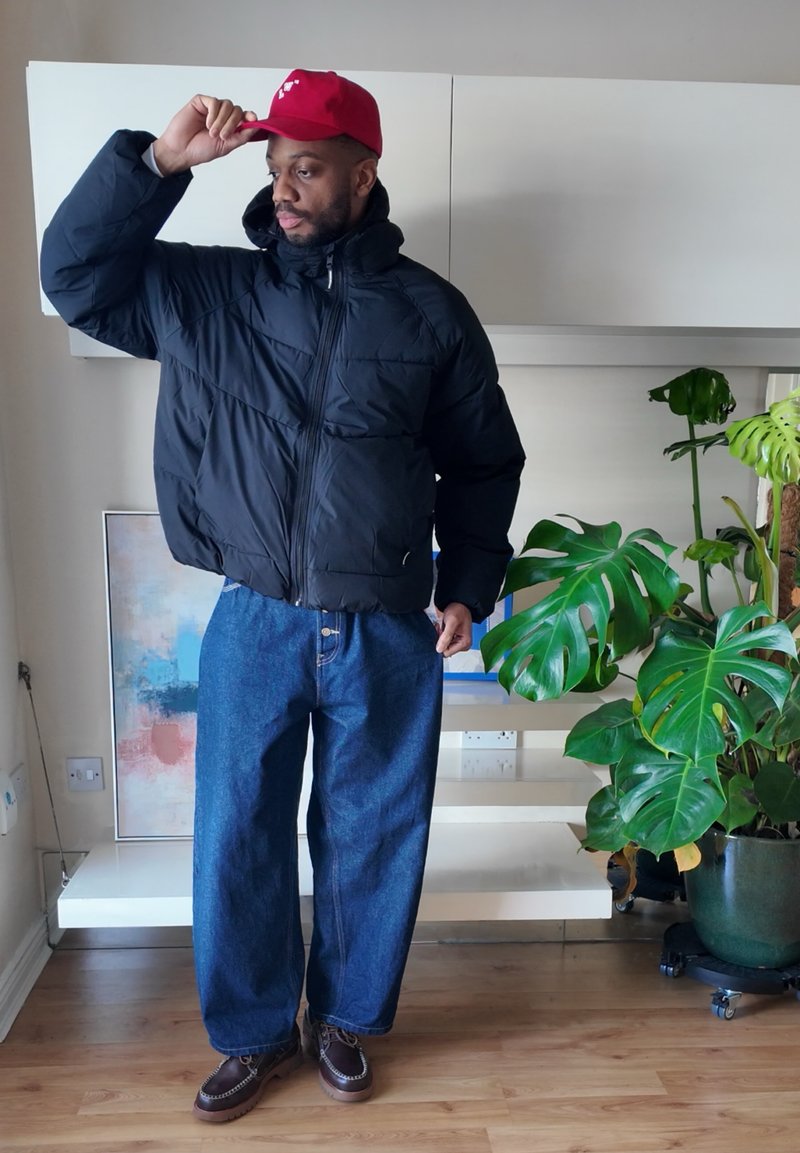Black puffer jacket, blue wide-leg denim jeans, brown shoes, and a red cap. Indoor setting with greenery and a modern backdrop.