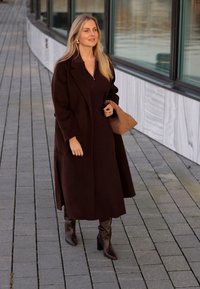 Μακρύ, σκούρο καφέ παλτό από υφασμένη ύφασμα, σε συνδυασμό με σκούρο καφέ εφαρμοστό φόρεμα και καφέ μπότες με τετράγωνη φτέρνα μέχρι το γόνατο. Περιλαμβάνεται πλεκτή τσάντα.