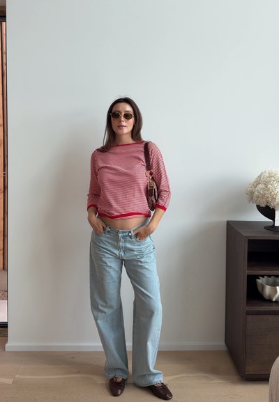Mujer con top de rayas rojas de manga larga, jeans azul claro, zapatos planos marrones, gafas de sol y llevando un bolso de hombro, de pie en una habitación cerca de un armario.
