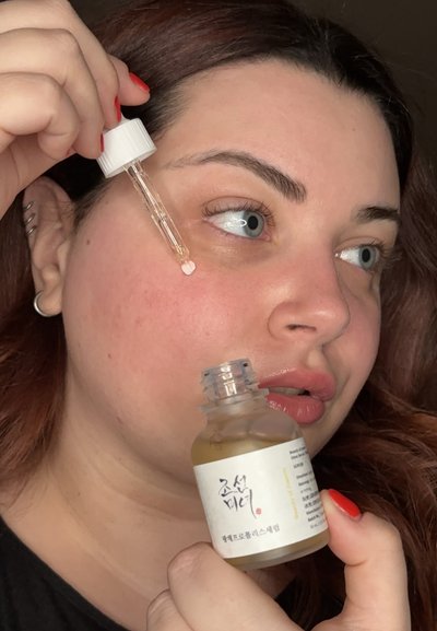 Botella transparente de suero con etiqueta blanca y texto negro, sostenida por una persona con cabello oscuro y uñas manicura roja; el gotero está ligeramente lleno.