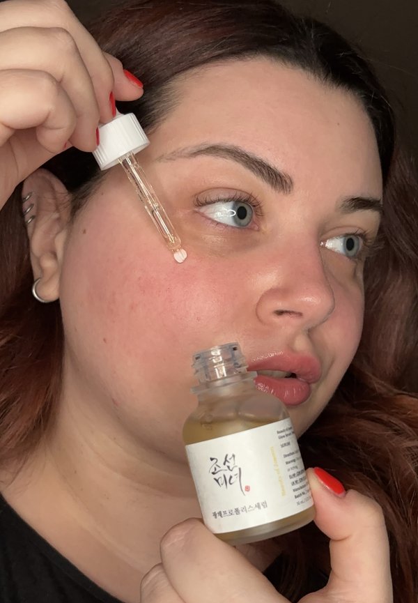 Bottiglia di siero trasparente con etichetta bianca e testo nero, tenuta da una persona con capelli scuri e unghie curate e rosse; il contagocce è leggermente riempito.