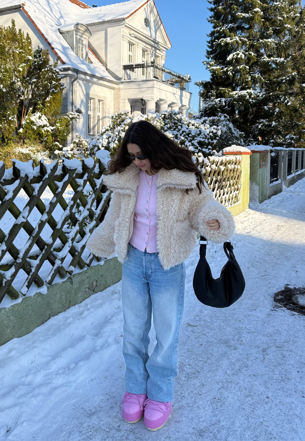 Femme en pull rose, veste beige en fausse fourrure, jeans bleus et chaussures roses, debout sur un trottoir enneigé, tenant un sac noir près d'une clôture en bois et d'une maison.