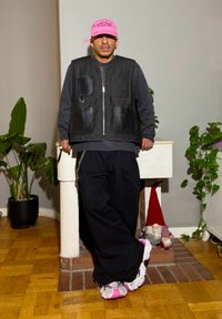 Veste en denim noir et cuir portée sur un haut gris, pantalons noirs amples et baskets blanches avec des accents roses. En arrière-plan, des plantes et des éléments décoratifs.