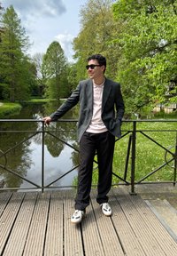 Uomo in un blazer grigio scuro doppiopetto sopra una camicia rosa chiaro, pantaloni neri e sneakers bianche, in piedi accanto a un fiume, circondato da vegetazione.