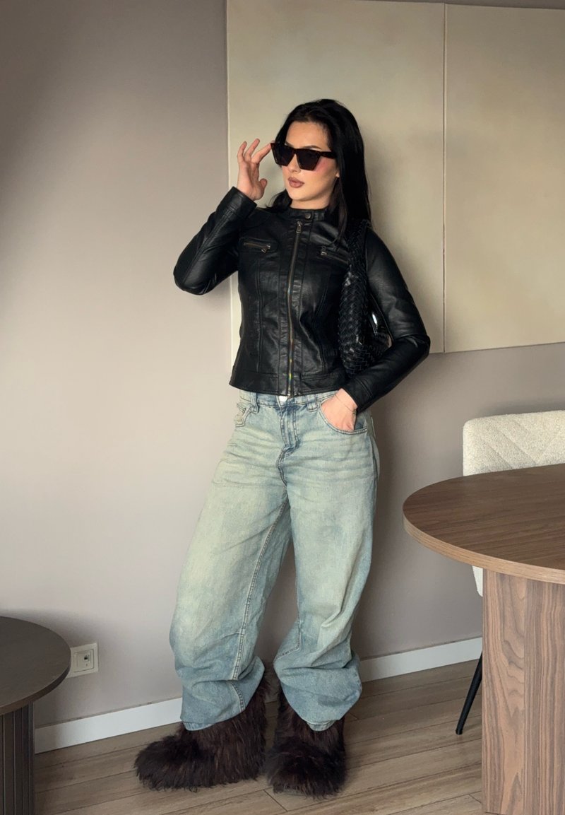 Femme portant une veste en cuir noire, un jean bleu clair surdimensionné, des bottes sombres en fourrure, des lunettes de soleil, et portant un sac à main noir texturé à l'intérieur.