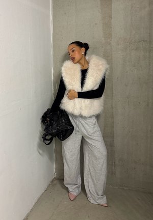 Mujer con chaleco blanco esponjoso, camiseta negra de manga larga, pantalones anchos grises, tacones rosas, sosteniendo un bolso negro tejido, de pie frente a paredes de concreto y blancas.