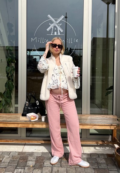 Chaleco de forro polar blanco, blusa con estampado, pantalones de pierna ancha rosa y zapatillas blancas. Bolso de mano negro. El fondo muestra cristal y vegetación.