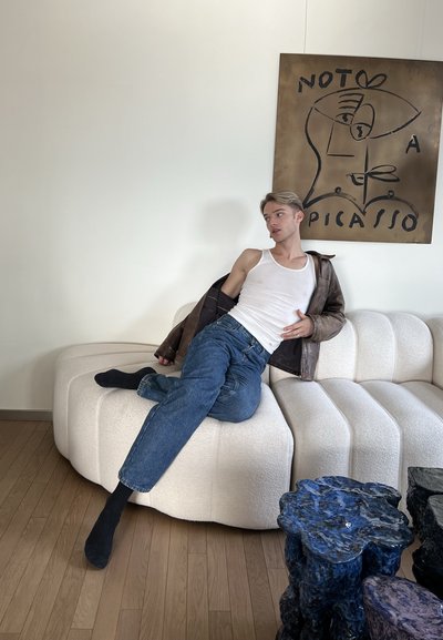 Joven con camiseta sin mangas blanca y chaqueta de cuero descansando en un sofá blanco con mesas cerámicas modernas azules y arte mural abstracto al estilo Picasso.