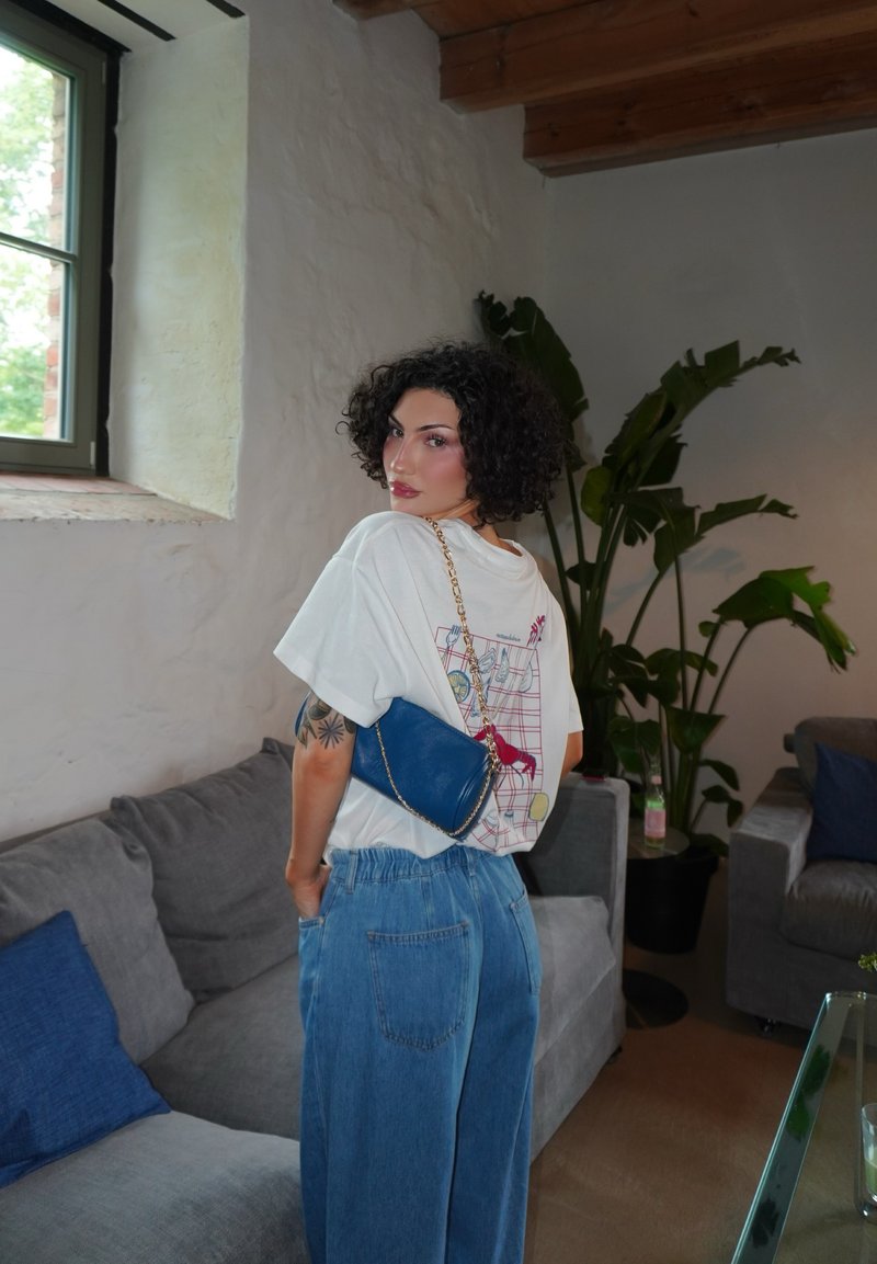 Femme aux cheveux noirs bouclés debout dans une pièce, portant un t-shirt graphique blanc et un jean bleu, avec un petit sac bleu à bandoulière chaîne.