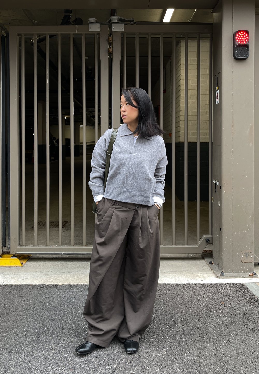 Harmaa neulepusero, jossa on kaulus, yhdistettynä väljien ruskeiden housujen kanssa. Henkilö seisoo aidatulla alueella, mustat kengät jalassa ja kantaa laukkua.