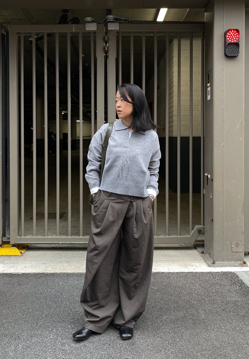 Pull gris en tricot avec col, associé à un pantalon marron ample. La personne se tient près d'une zone grillagée, portant des chaussures noires et portant un sac.